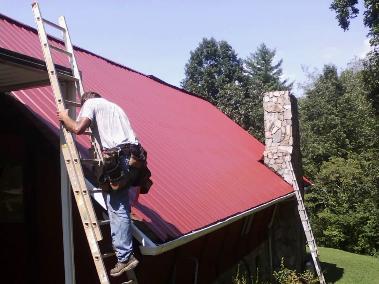 Roofer on a ladder beside a red metal roof and stone chimney — Soffit & Fascia Repair by Roofing Brothers in Weimar